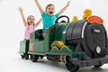 Two happy children riding a green electric Classic Railroad ride-on train toy with their arms raised in excitement. The train is trackless so it can ride anywhere.